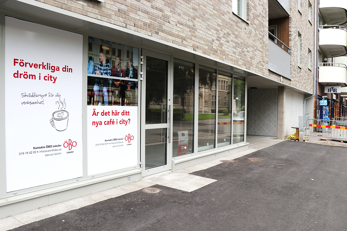 Husfasad med stora skyltfönster i bottenplan och flera våningar med balkonger ovanför. På fönstren syns affischer med text från ÖBO Lokaler.