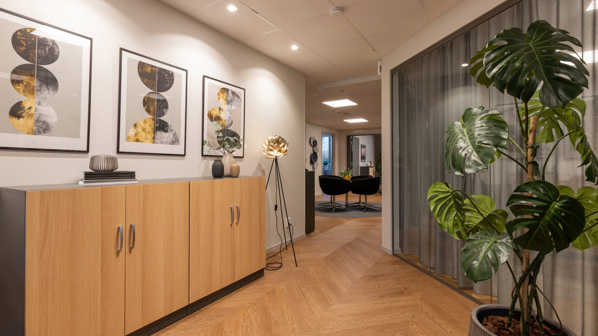 Korridor med lång träskänk och tre inramade tavlor på väggen; till höger finns en glasvägg med gardin och en stor krukväxt, och längre bort syns sittgrupp.