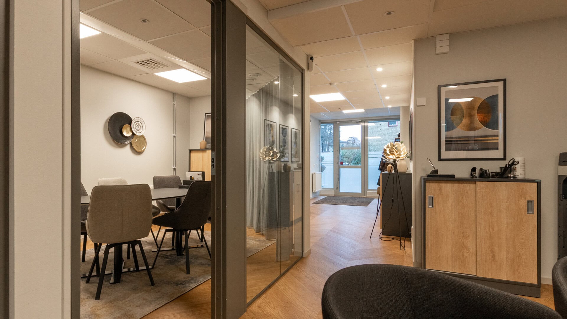 Korridor med glasväggar och ett mötesrum till vänster med bord och flera stolar; längre bort syns entrédörr.