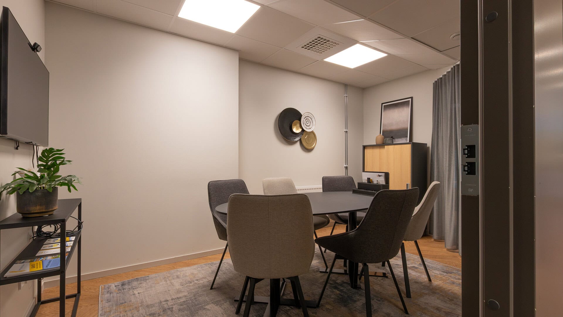 Mötesrum med ovalt bord och sex stoppade stolar; en TV på väggen, en växt på ett sidobord och väggdekoration samt förvaringsskåp i bakgrunden.