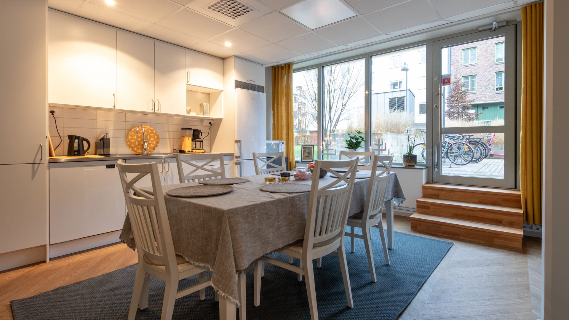 Kök med lång bänkskiva och vit skåpsinredning; matbord med vita stolar framför stora fönster och en glasdörr som leder ut, med trappa upp till dörren