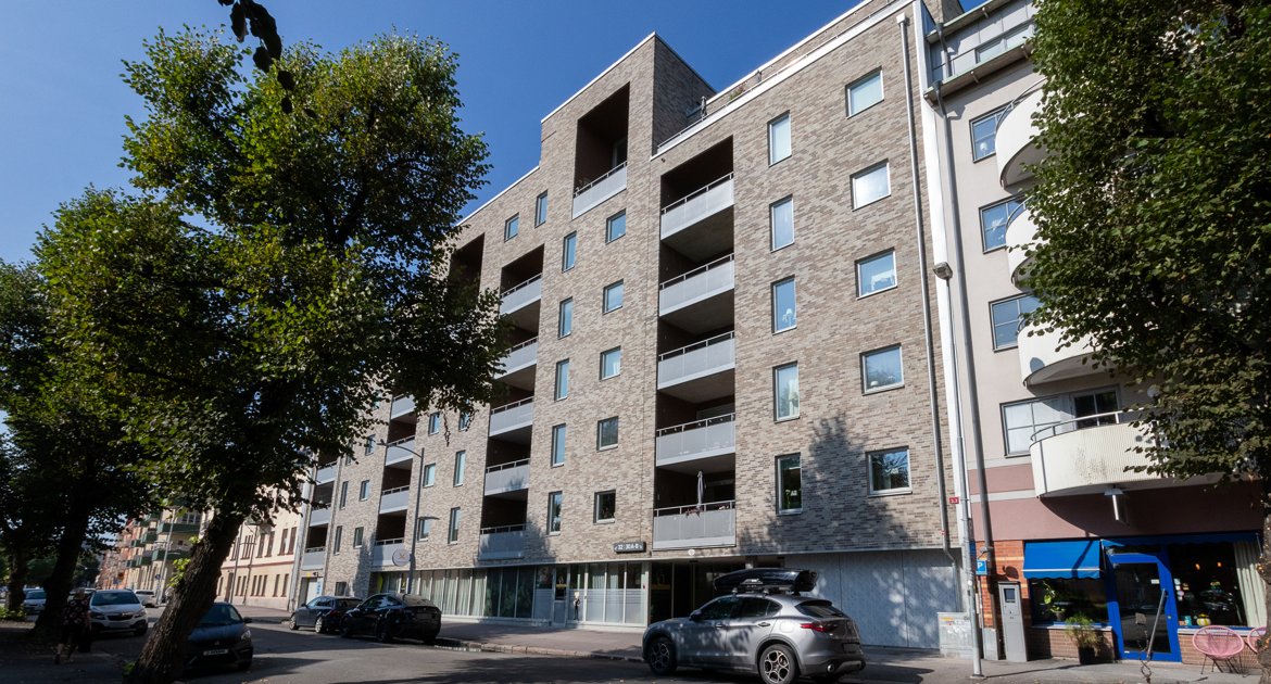 Gata med flerbostadshus i flera våningar till höger. Husen har balkonger och ljus tegelfasad. Parkerade bilar och träd syns längs gatan.