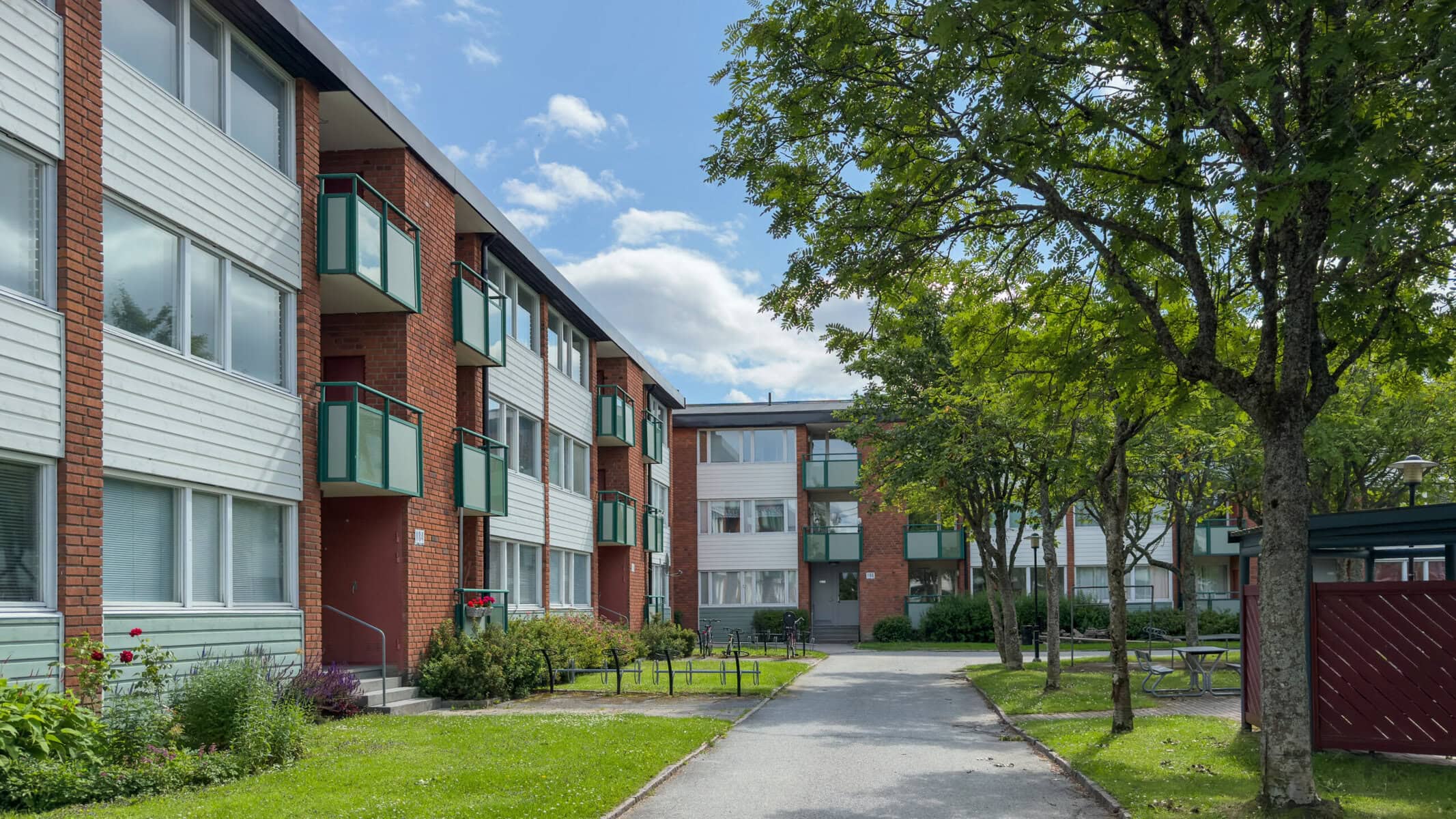 Gångväg mellan flerbostadshus med balkonger, träd, gräsytor och gemensamma sittplatser.