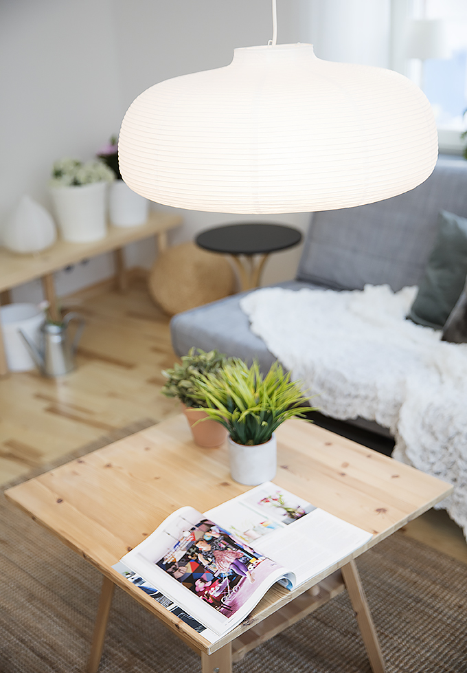 Vardagsrum med soffa, soffbord och en lampa. På soffbordet finns en uppslagen tidning och en blomma.