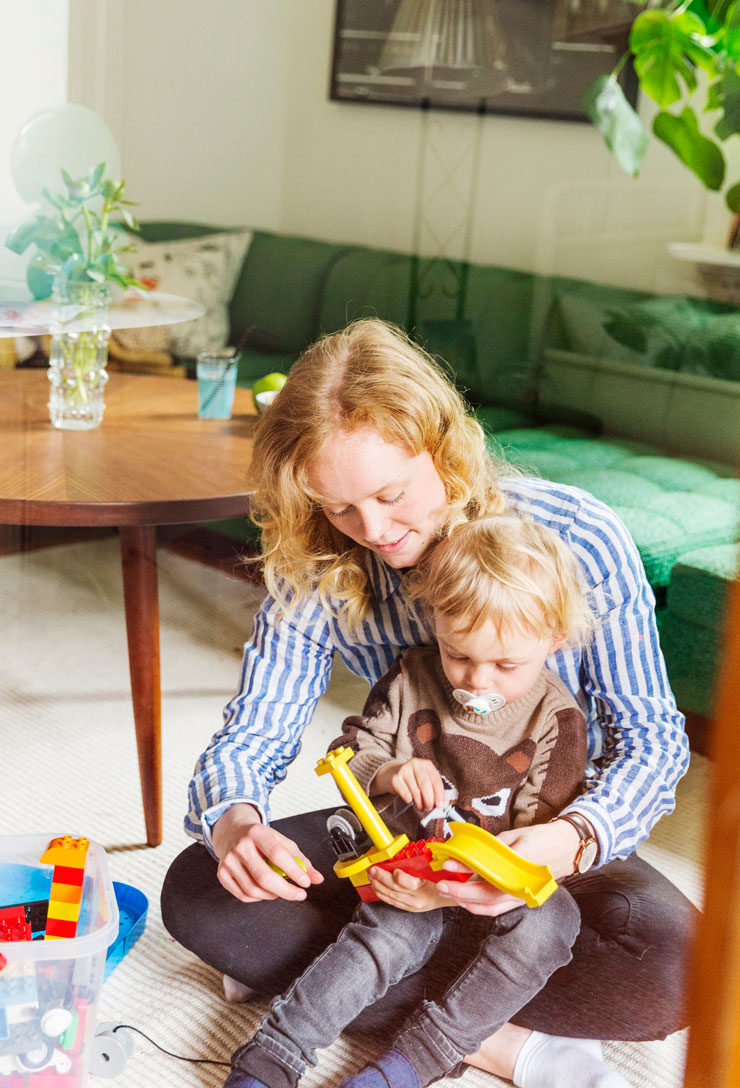 Förälder sitter på golvet framför en grön soffa. I knät sitter barnet och de leker med duplo.