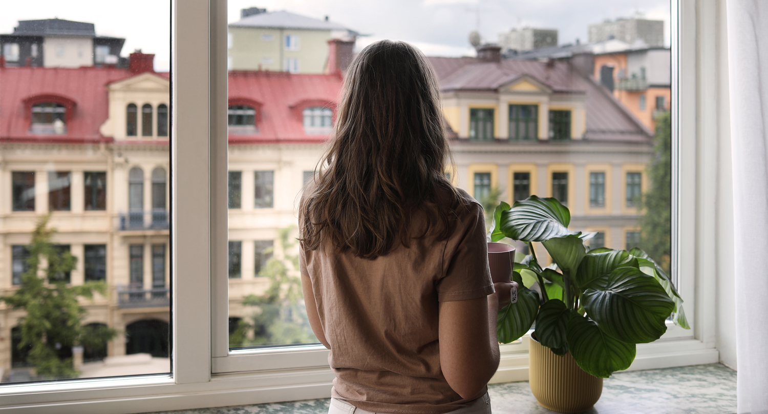 Kvinna med beige tröja och långt hår blickar ut över Stortorget från fönster i lägenhet i kvarteret Hållstugan.