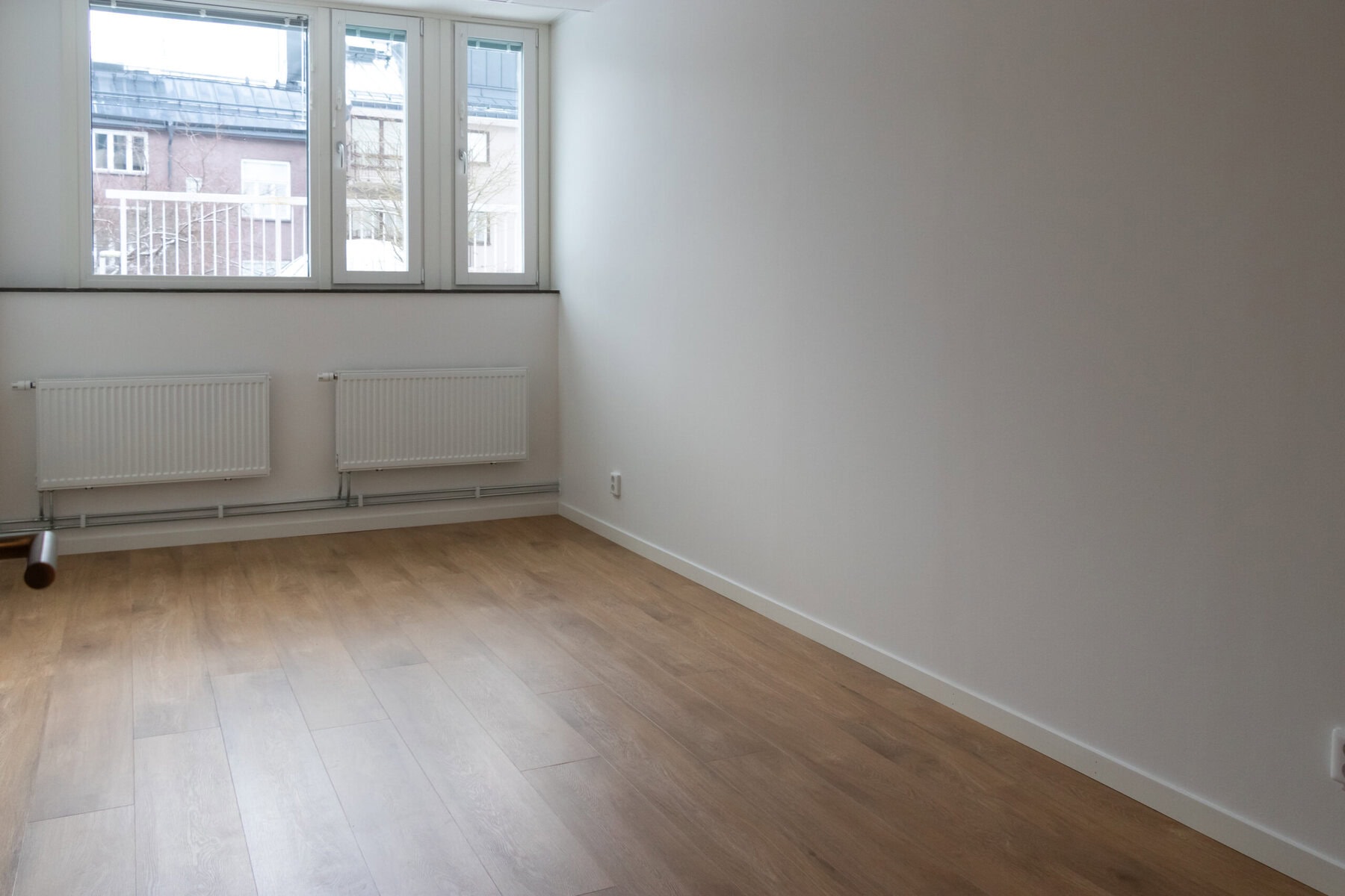 Tomt rum med vita väggar och parkettgolv. Ett stort fönster mot innergården.