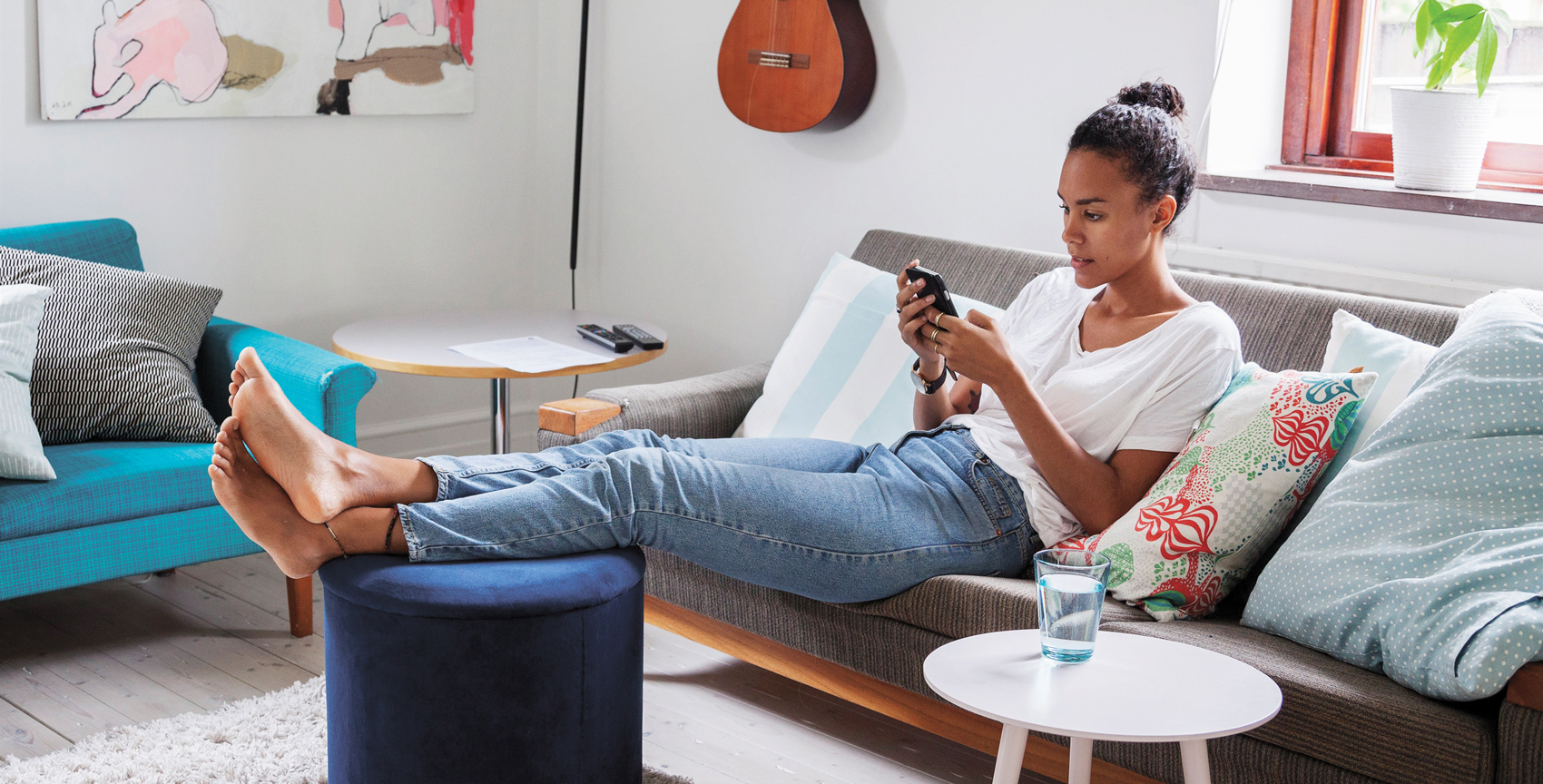 Kvinna i tjugoårsåldern sitter i soffan med benen på en puff och läser på sin mobiltelefon.