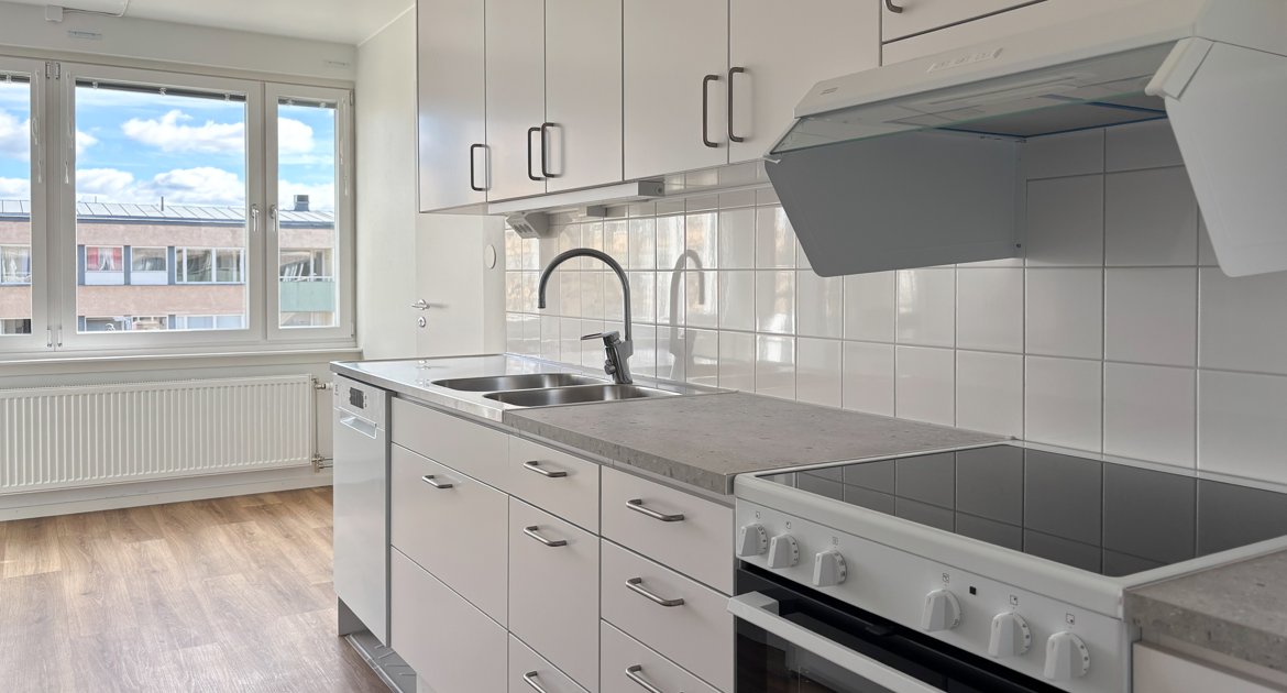 Kök med vita skåpluckor, grå bänkskiva, spis, diskmaskin och matplats vid fönster.