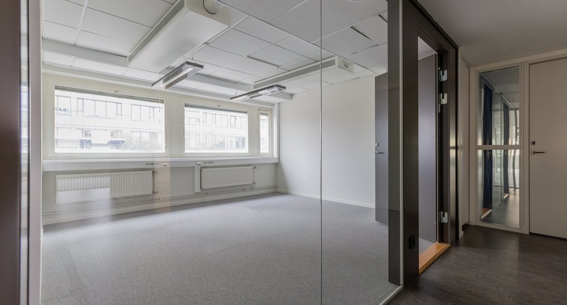 Kontorsrum med glasvägg och dörr. Grå matta, stora fönster med element under och vy mot korridor med flera dörrar.