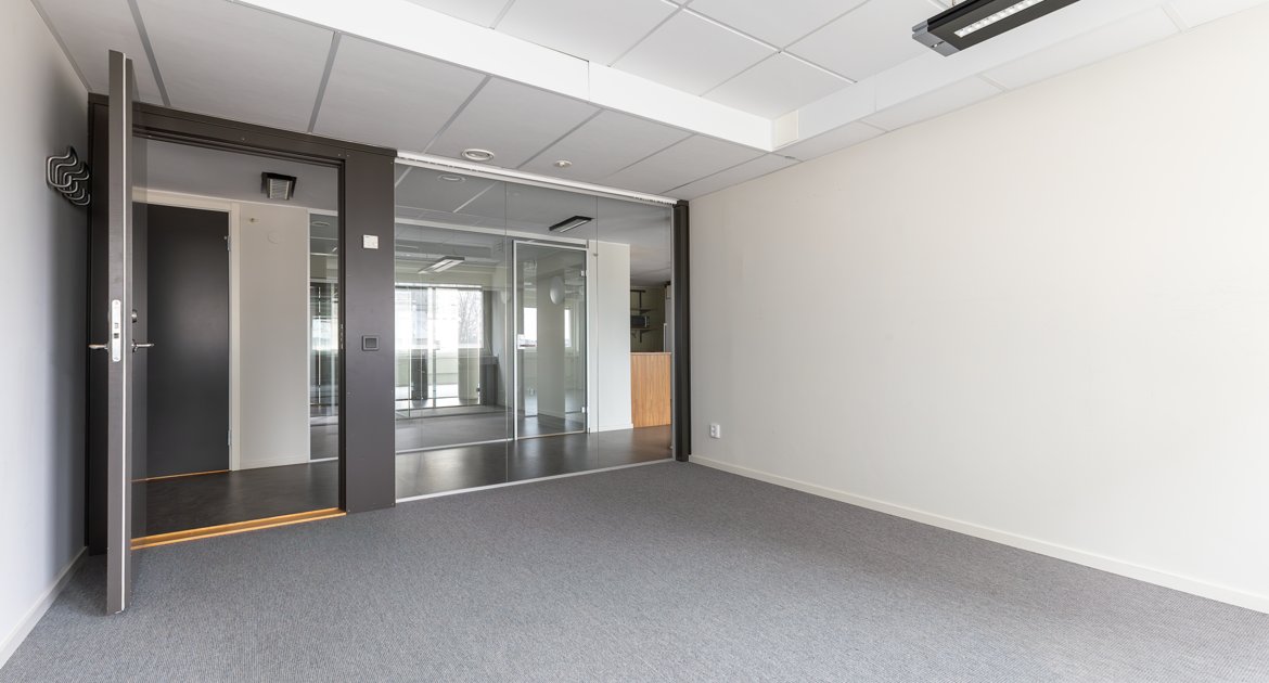 Kontorsrum med glasväggar och dörrar. Grå matta, ljusa väggar och utsikt mot angränsande rum genom glaspartier.