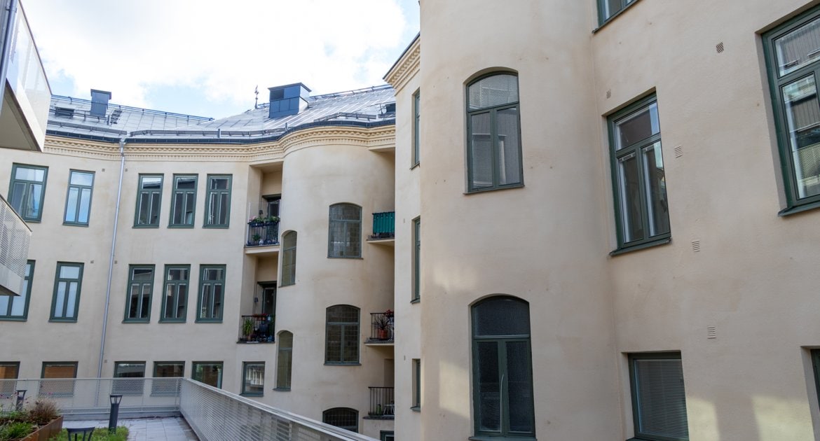 Innergård med ljus putsad bostadsfasad i flera våningar, gröna fönsterkarmar och små balkonger med växter.