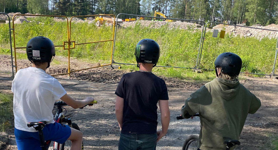 Tre pojkar i cykelhjälmar tittar ut över en obebyggd yta med grus, gräs och en grävmaskin.