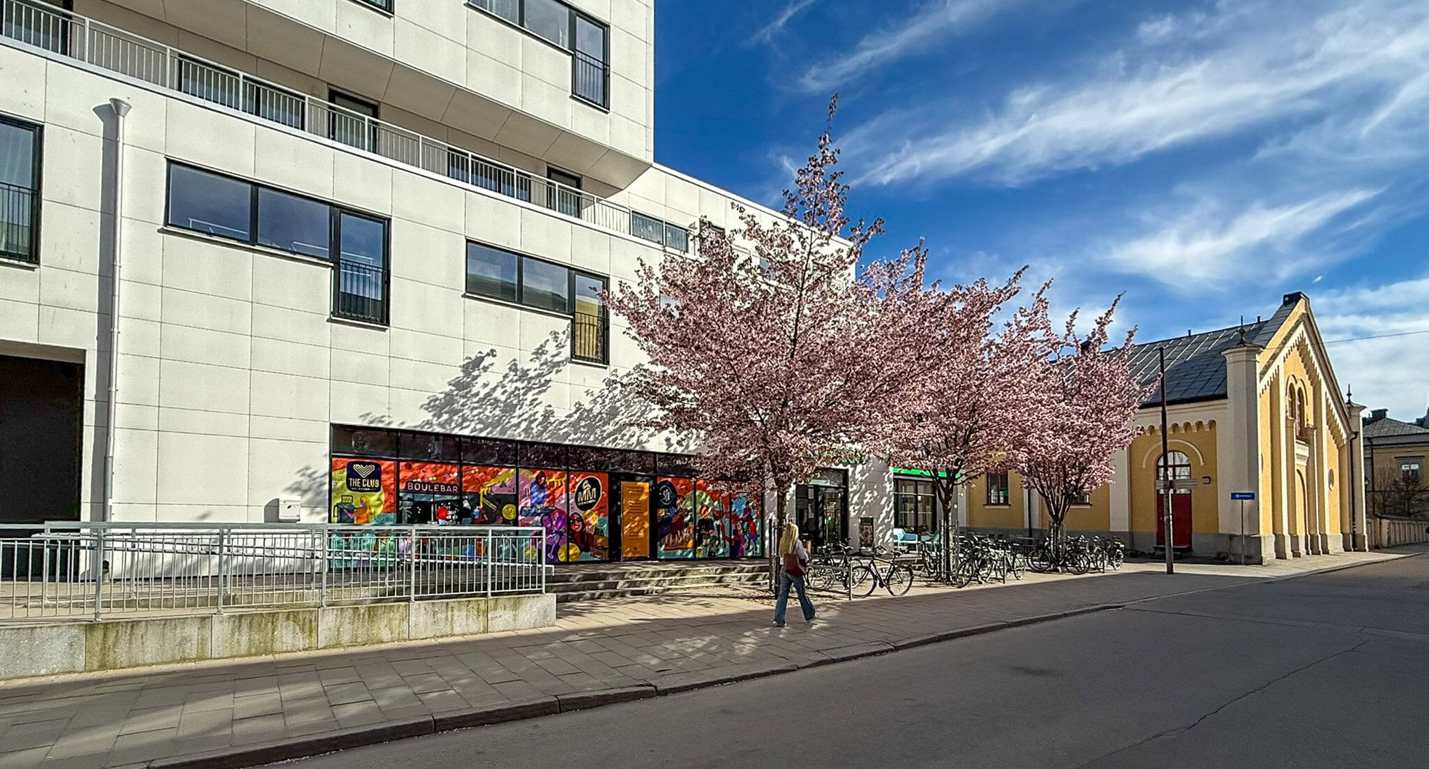 Solbelyst stadsgata med blommande körsbärsträd framför ett flerbostadshus och butikslokaler. En person promenerar längs trottoaren bredvid parkerade cyklar.