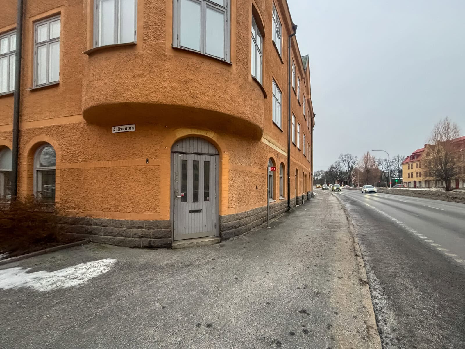 Gatuvy längs byggnadens fasad med entrédörr och trottoar, bred väg intill och bilar i bakgrunden.