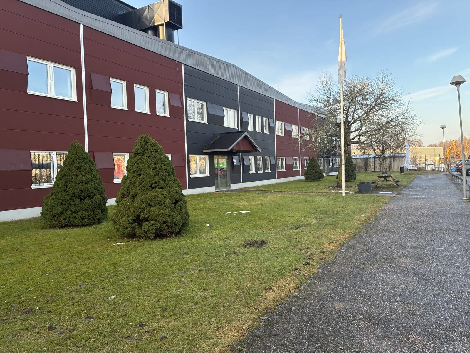Industrifastighet i rött och grått med entréparti, gräsyta med buskar och flaggstång samt gångväg längs byggnaden.