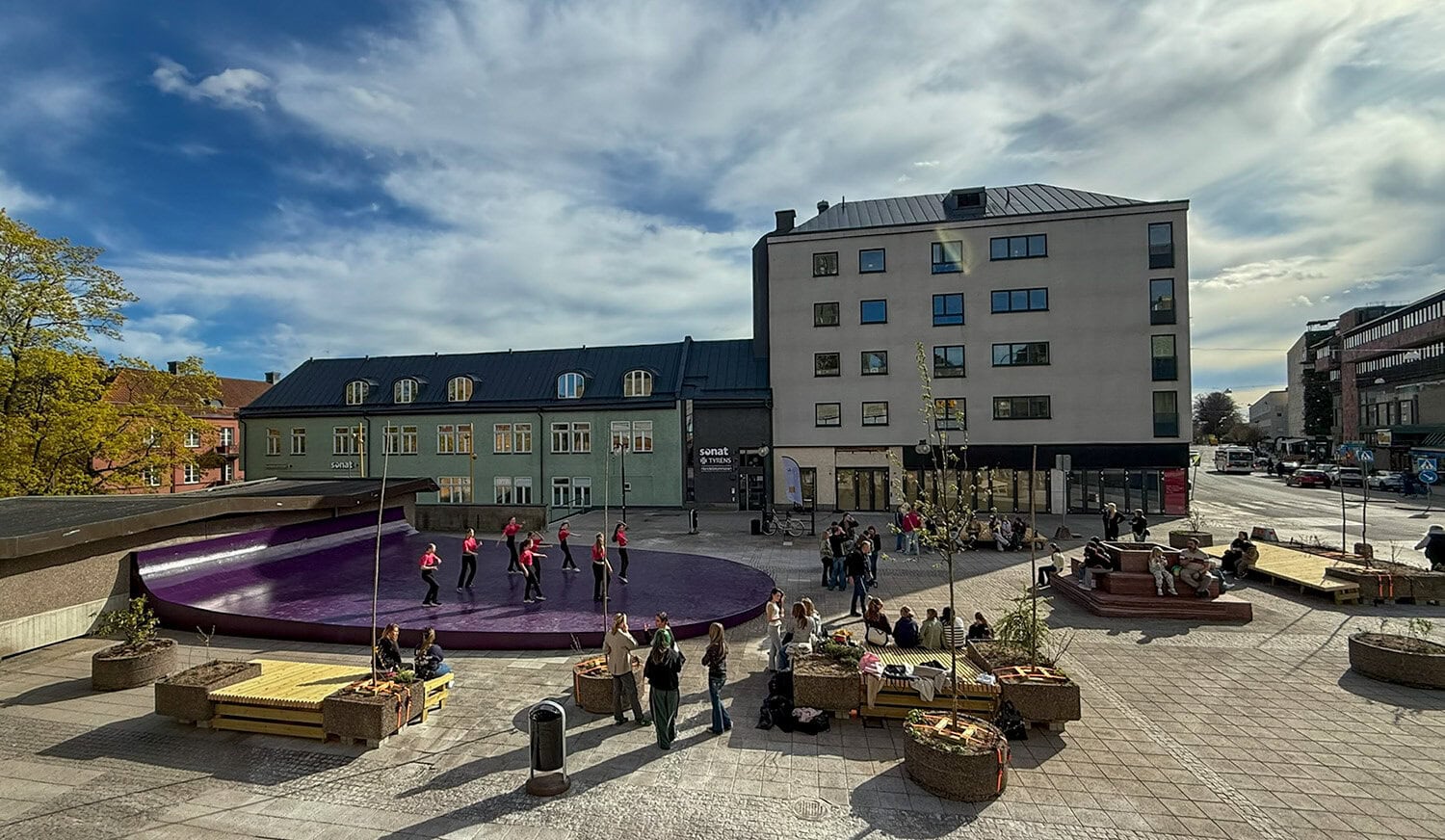Ett öppet torg med en stor lila dansscen där en grupp personer uppträder inför publik. Runt scenen sitter och står människor på bänkar och i små grupper. I bakgrunden syns flerbostadshus, träd och himmel med stora moln.