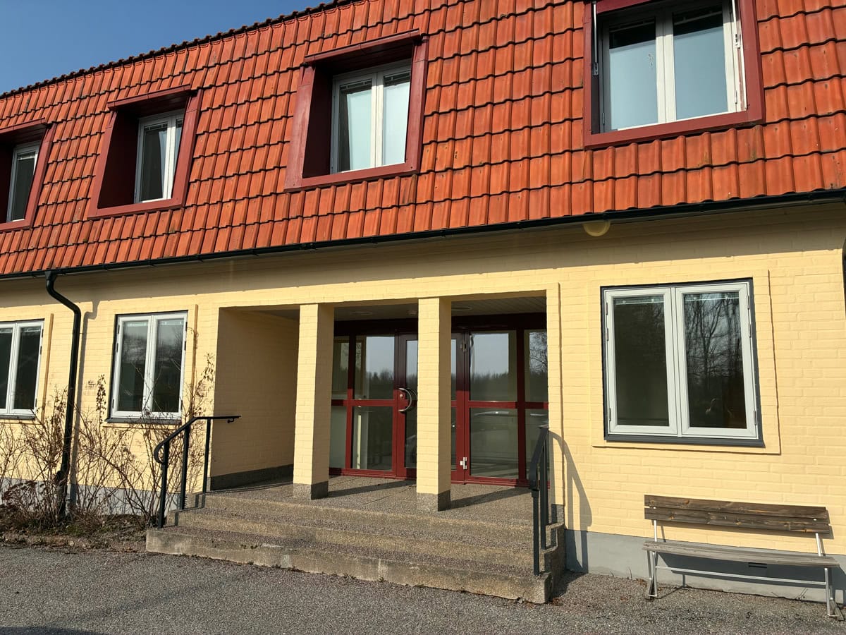 Entré till gul tegelbyggnad med rött tegeltak, trappa och bänk utanför.