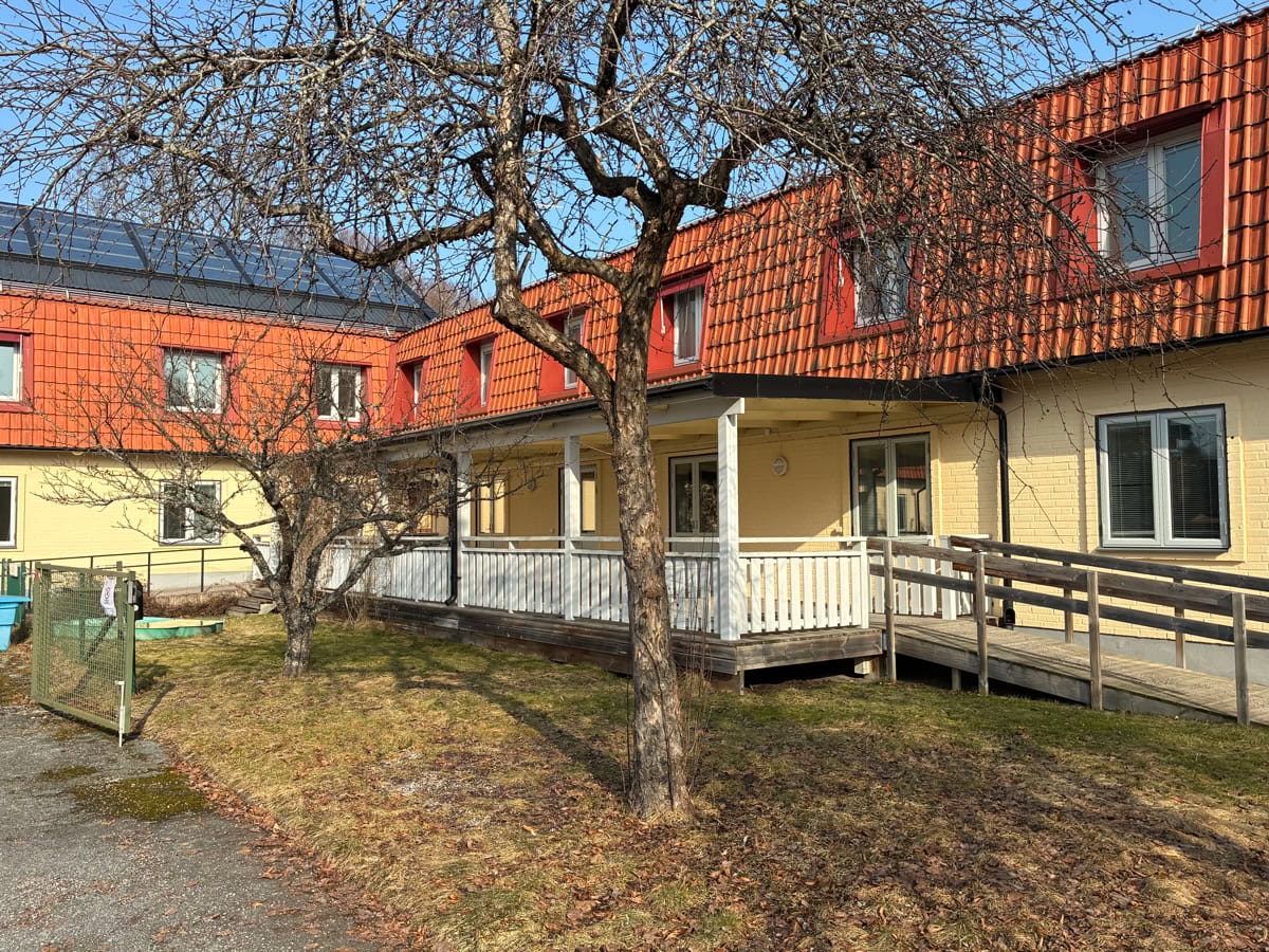Gul tegelbyggnad med rött tegeltak, veranda och tillgänglighetsramp, omgiven av träd och gräsyta.