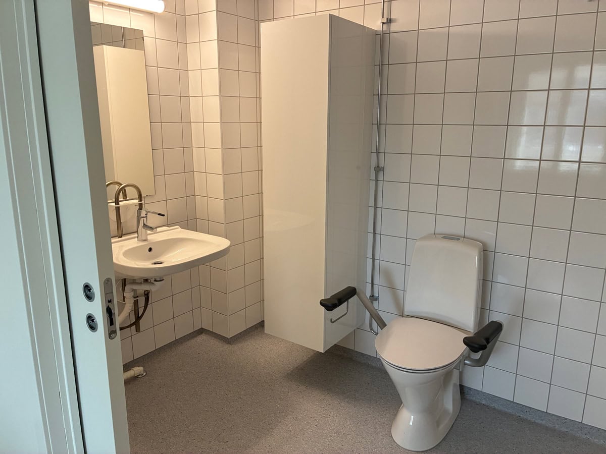 Tillgänglighetsanpassat badrum med toalett med armstöd, handfat och vita kakelväggar.